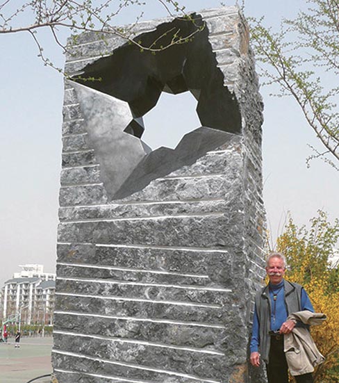 A smiling Jon Hudson leans against his “Synchronicity Beijing.” This commision was carved in 2011 to commemorate the 100th anniversary of Tsinghai University, Beijing, China, founded with assistance from the U.S.A.. Erected on the campus, it is a towering 16 feet tall, 8 feet wide and 2 feet deep.