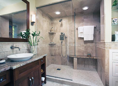 Warm-colored marble, natural granite tones, a maple vanity, and an open airy shower and drying area are at the heart of the well-composed welcoming space of this in-home luxury spa. With towels and shampoo close at hand and a bench outside the shower that provides a convenient spot to towel off, the client is sure to feel pampered in the new bathroom. A cabinet, shelf space, and a vanity containing drawers combine to provide stylish storage, while a frameless glass shower with light-hued marble tile and flooring gives an expansive appearance to a somewhat limited space.  Designer: John Sylvestre, CKD; Photo ©Karen Melvin Photography