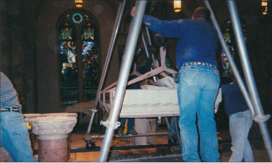 Step 8: After getting the piece into position in front of the altar and laying the piece down in the horizontal position, a rolling A-frame with a 14-inch aluminum I-beam and chainfalls were used to hoist the piece high enough to come over the top of the waiting base of four columns..Look closely and you will see that the wheels are being kept straight by a track made out of channel irons. Its important to remember this technique, as carrying such a heavy load on an A-frame support is very hard to manage and unwieldy. This task marks the end of day one of installation.