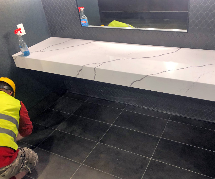 A complete final cleanup is standard practice for StoneMark installations, Including large commercial jobs. Here, a StoneMark installer works on the tiled floor under a mitered vanity.  