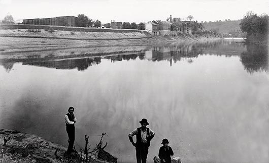 This archive photo shows a riverside view of Buffalo Trace Distillery, circa late 1800s. Visible on the left: the tall, brick sides of the three-story warehouses rise above the other buildings