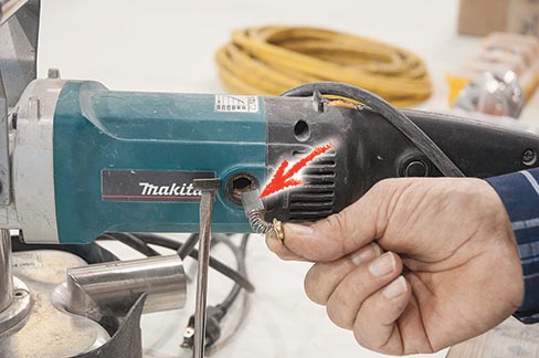 Carbon brush locations on a Makita (NEED MODEL#) lie behind easily removed buttons located just in front of the handle. Be careful–they are spring retained, as shown in the above photo. This model is attached to a Scarab machine; it’s not necessary to remove the cover to access the brushes.